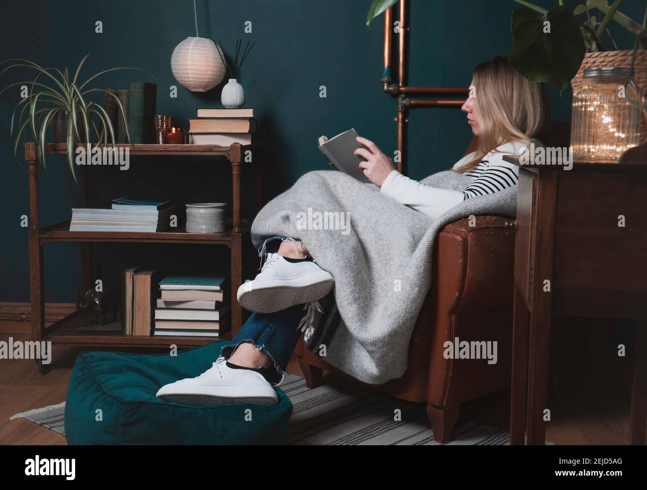 Une jeune femme caucasienne à la maison s'est emmêlée dans une couverture sur une chaise en cuir dans la salle de séjour ou a étudié la lecture d'un livre. Atmosphère confortable et détendue avec le contemplaa Banque D'Images