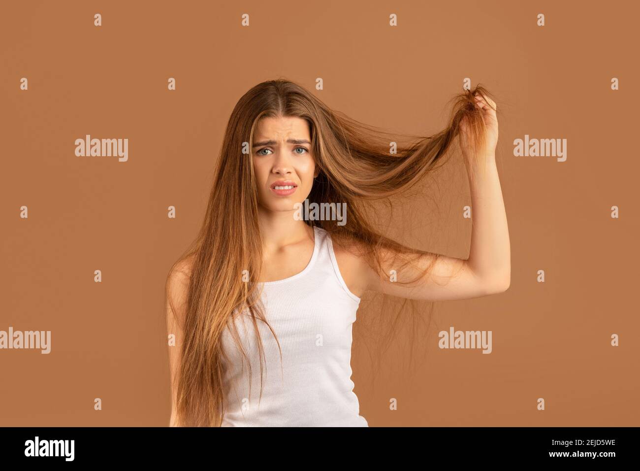 Femme millénaire frustrée montrant ses longs cheveux abîmés sur fond de studio brun. Concept de services de coiffure Banque D'Images