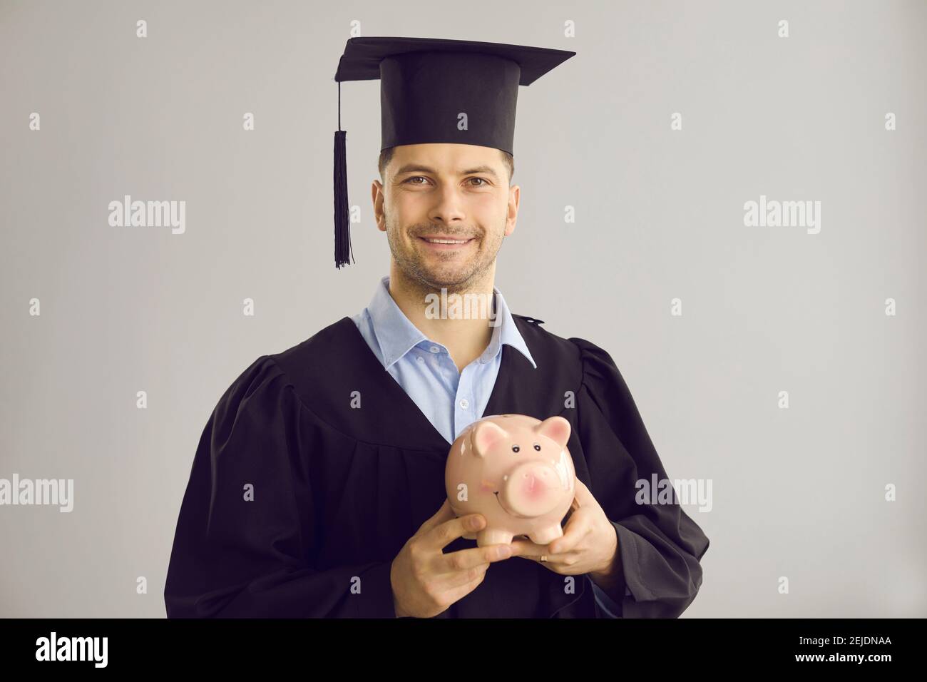 Joyeux diplômé en chapeau et robe tenant la banque de porc avec de l'argent pour l'éducation de troisième cycle Banque D'Images