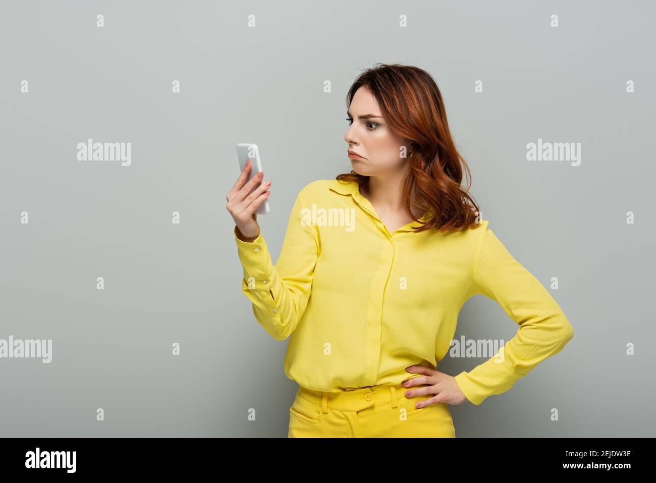 choqué femme regardant le téléphone mobile en se tenant debout avec la main sur la hanche sur le gris Banque D'Images
