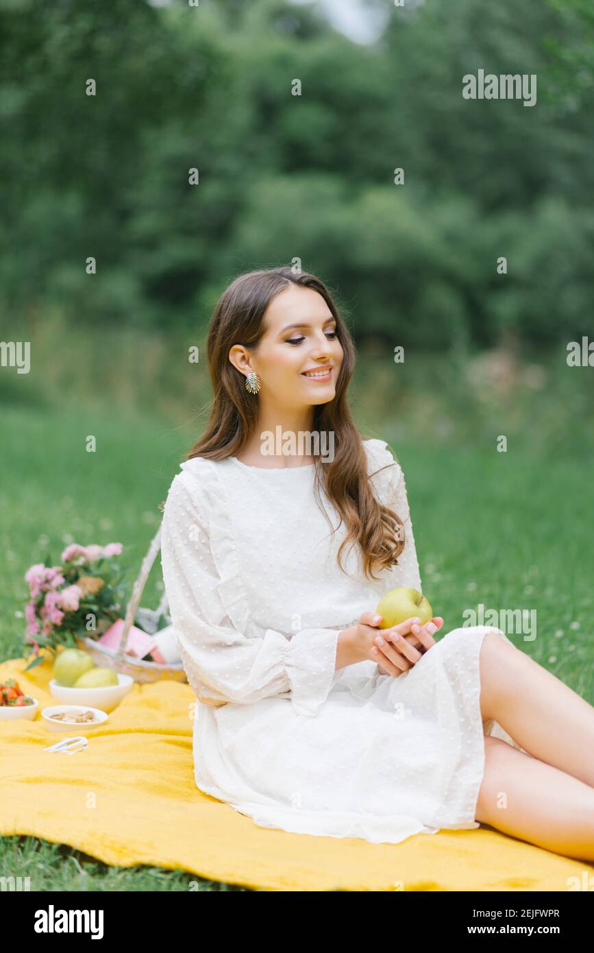Jeune fille caucasienne souriante assise à l'extérieur. Jeune femme artistique créative portant des vêtements décontractés ayant un pique-nique avec pomme le jour d'été. Banque D'Images
