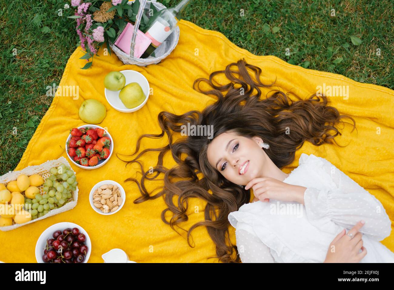 Souriante jeune fille caucasienne se trouve sur le dos à l'extérieur. Vue de dessus. Jeune femme artistique créative portant des vêtements décontractés ayant un pique-nique avec des fruits sur su Banque D'Images