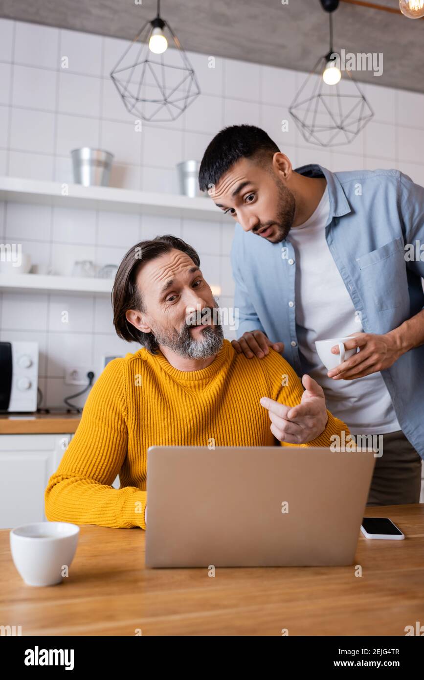un homme stupéfait qui pointe vers un ordinateur portable près d'un fils hispanique dans la cuisine Banque D'Images