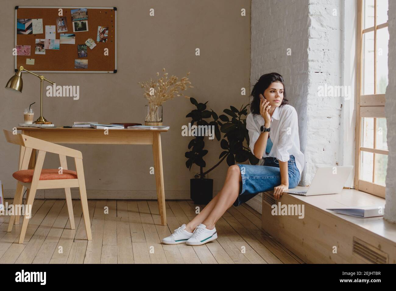 Jeune belle femme en tenue décontractée communiquant sur un smartphone assis sur un rebord de fenêtre dans un bureau moderne. Banque D'Images