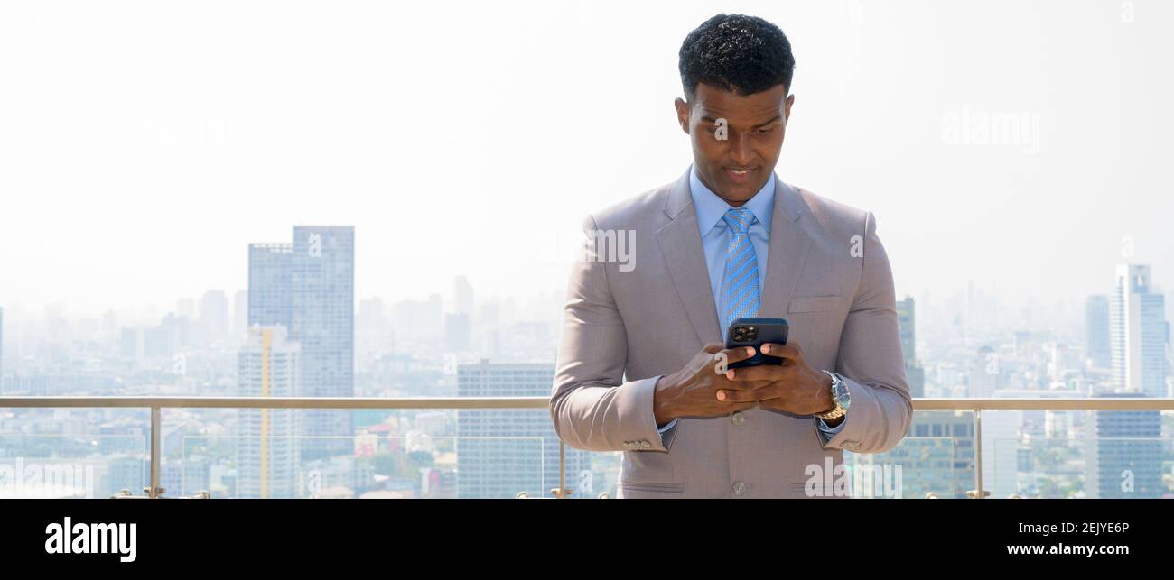 Portrait d'un beau jeune homme d'affaires africain vêtu de son costume tout en souriant et à l'aide d'un téléphone portable Banque D'Images