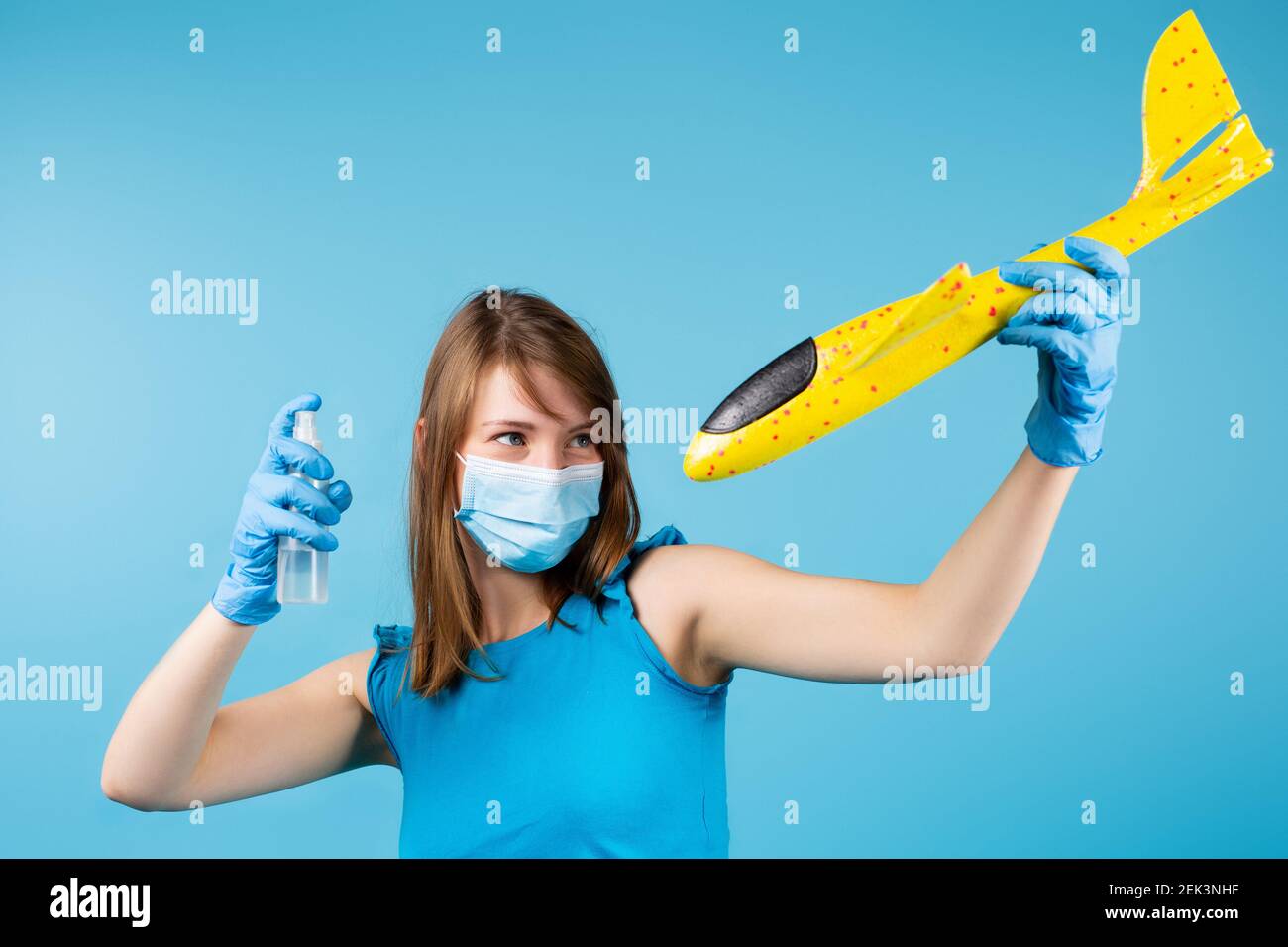 Une fille dans un masque médical et des gants lave le spray antiseptique sur un avion jaune de jouet sur un fond bleu. Lutter contre la propagation du coronavirus. Banque D'Images