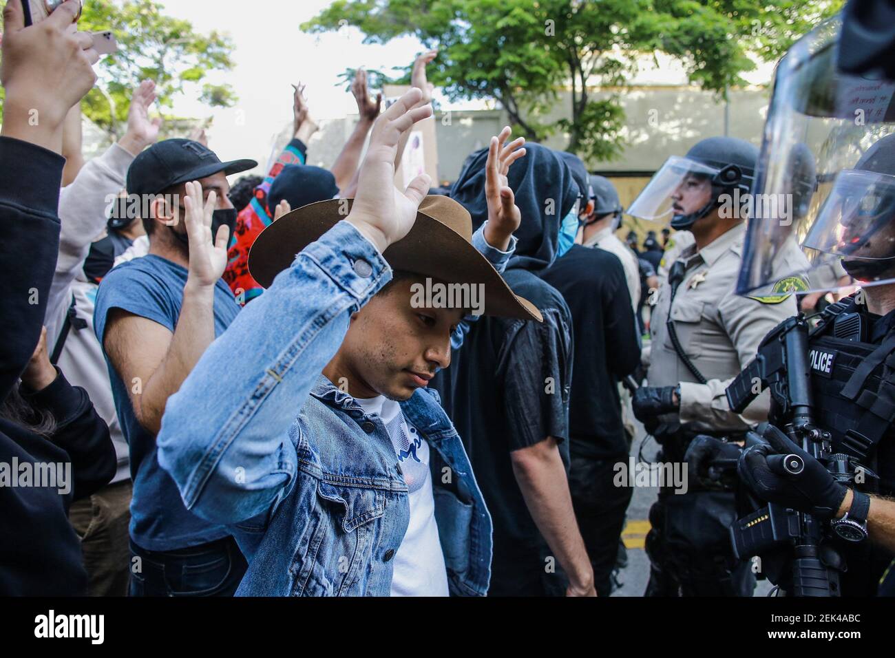Un manifestant portant un chapeau de cow-boy et une veste jean se tient  devant une ligne de gendarme, les mains levées dans les airs, tandis que  les manifestants scandent, « les mains