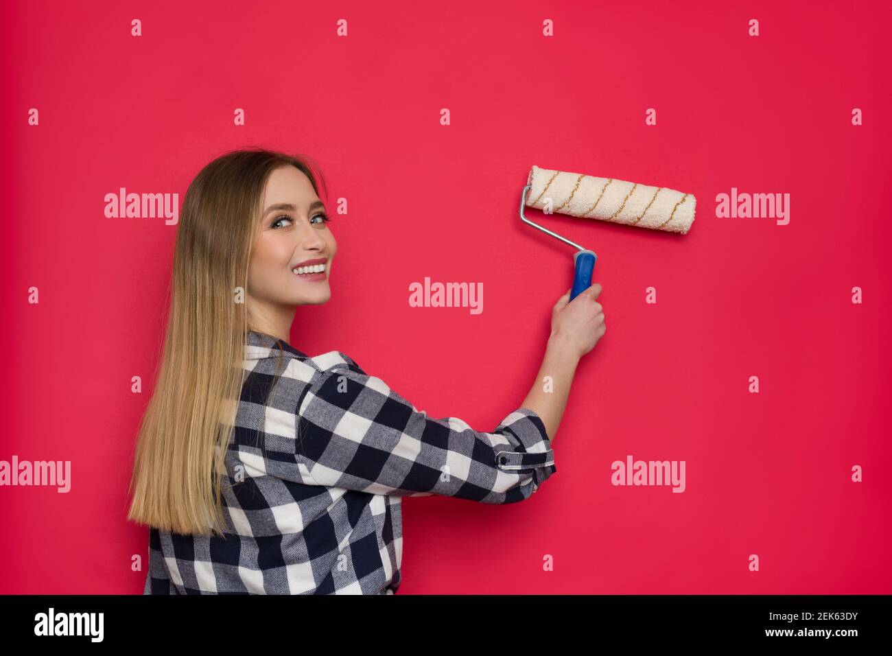 Souriante jeune femme blonde dans la chemise de bûcheron peint le mur rouge avec le rouleau de peinture et regardant la caméra au-dessus de l'épaule. Taille haute. Banque D'Images