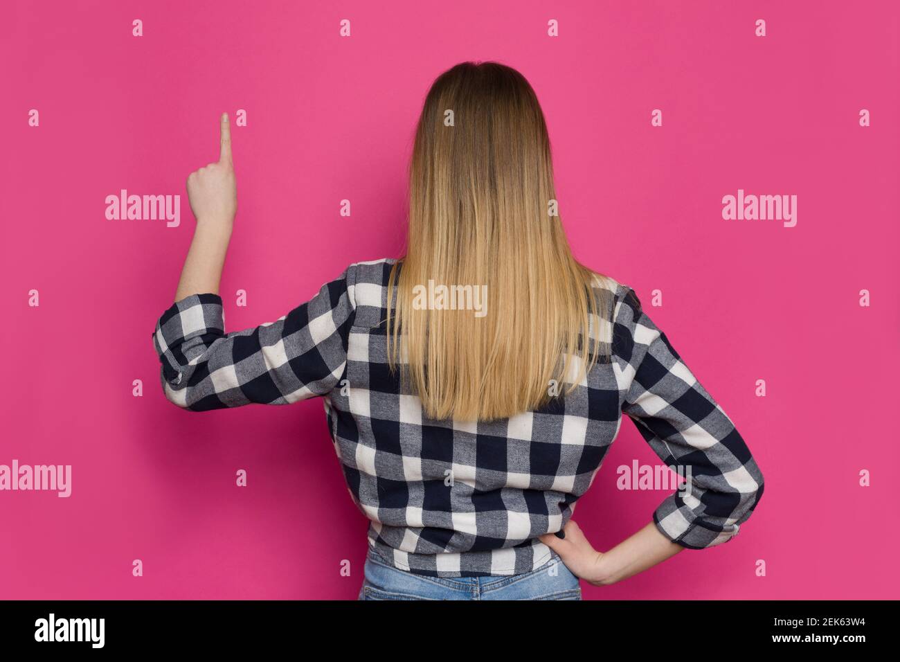 Une jeune femme en chemise de bûcherons pointe vers le haut. Vue arrière. Taille haute studio tourné sur fond rose. Banque D'Images