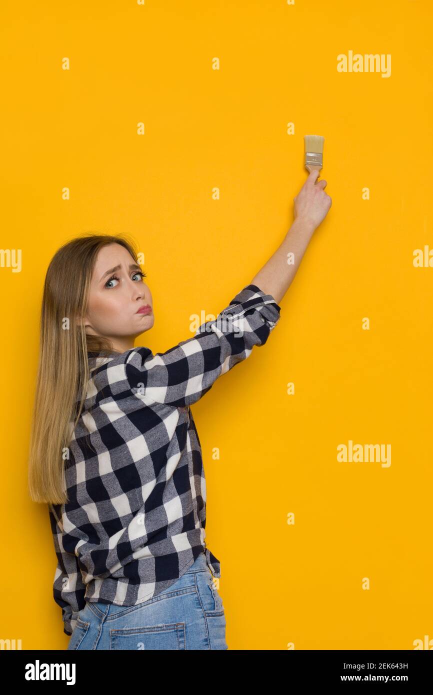 Inquiète jeune femme blonde dans la chemise et le Jean de bûcheron peint le mur jaune avec la brosse et regardant l'appareil photo au-dessus de l'épaule. Taille haute. Banque D'Images
