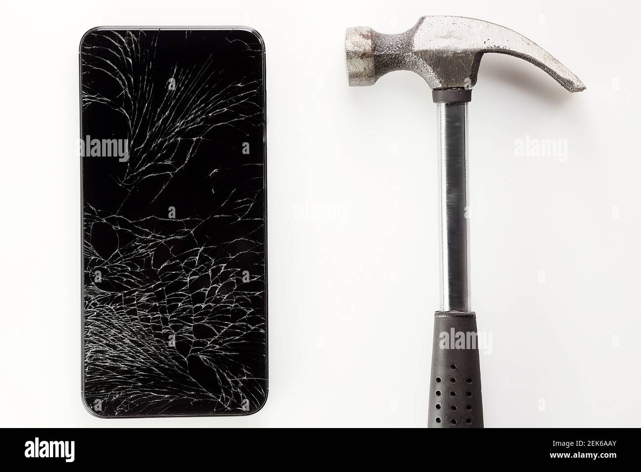 Un smartphone noir cassé avec un écran fissuré et un marteau en métal se trouve sur un fond blanc. Banque D'Images