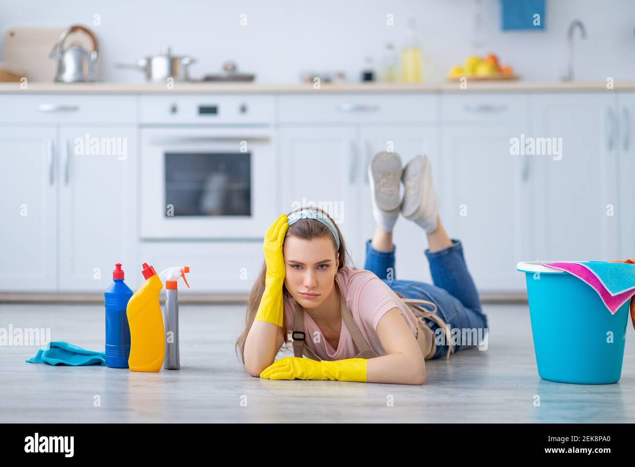 Une jeune femme fatiguée, couchée sur le sol parmi les fournitures de nettoyage, épuisée des travaux ménagers dans la cuisine, l'espace de copie Banque D'Images