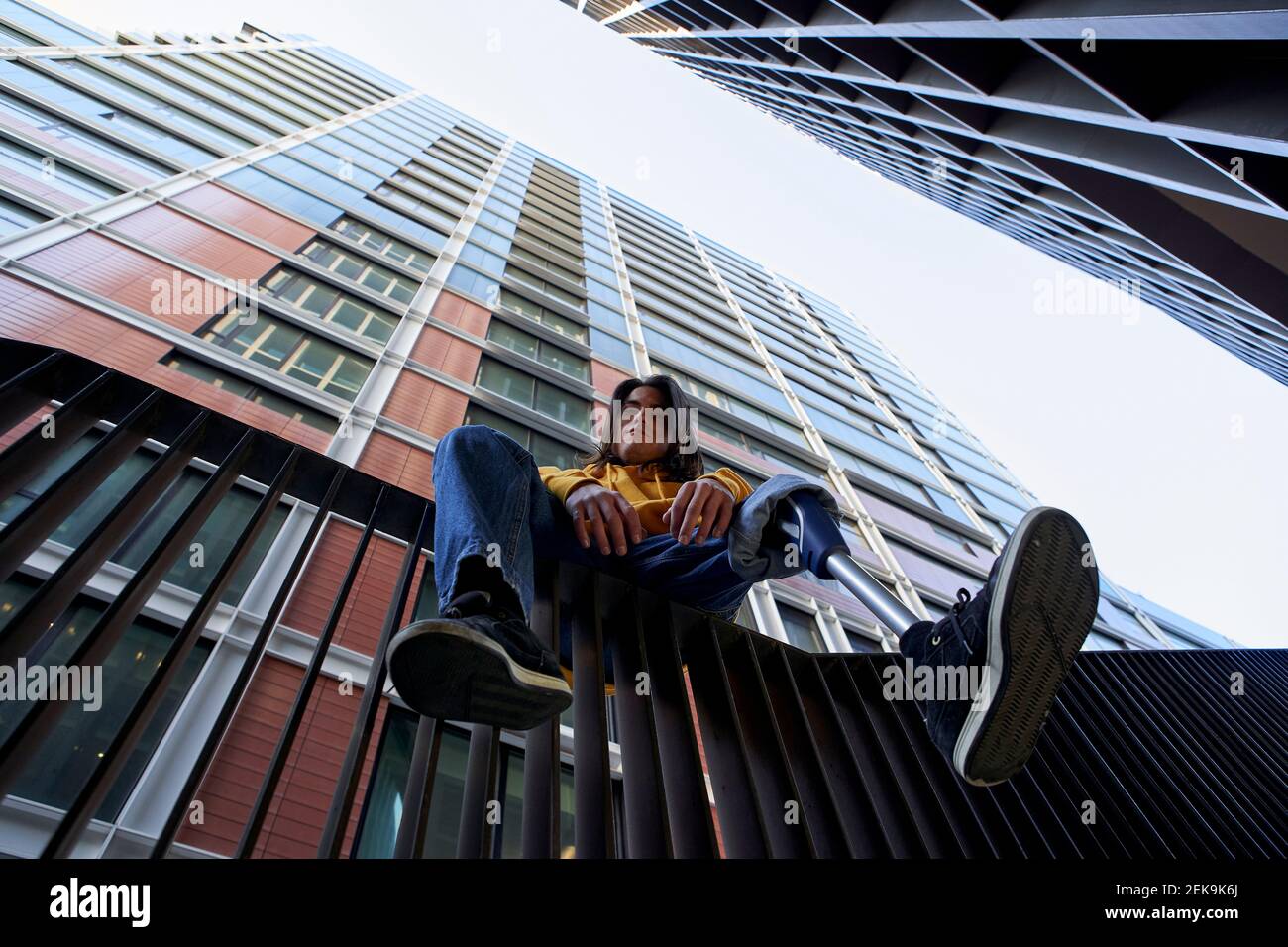 Jeune homme handicapé avec prothèse de jambe assis sur une main courante métallique contre le gratte-ciel Banque D'Images