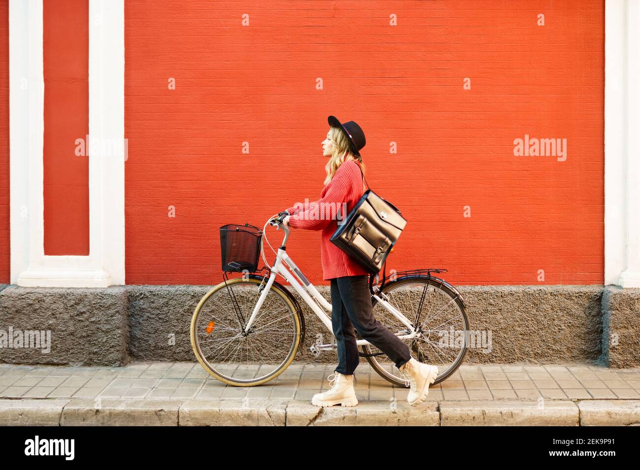 Jeune femme portant un chapeau poussant un vélo devant le mur rouge Banque D'Images
