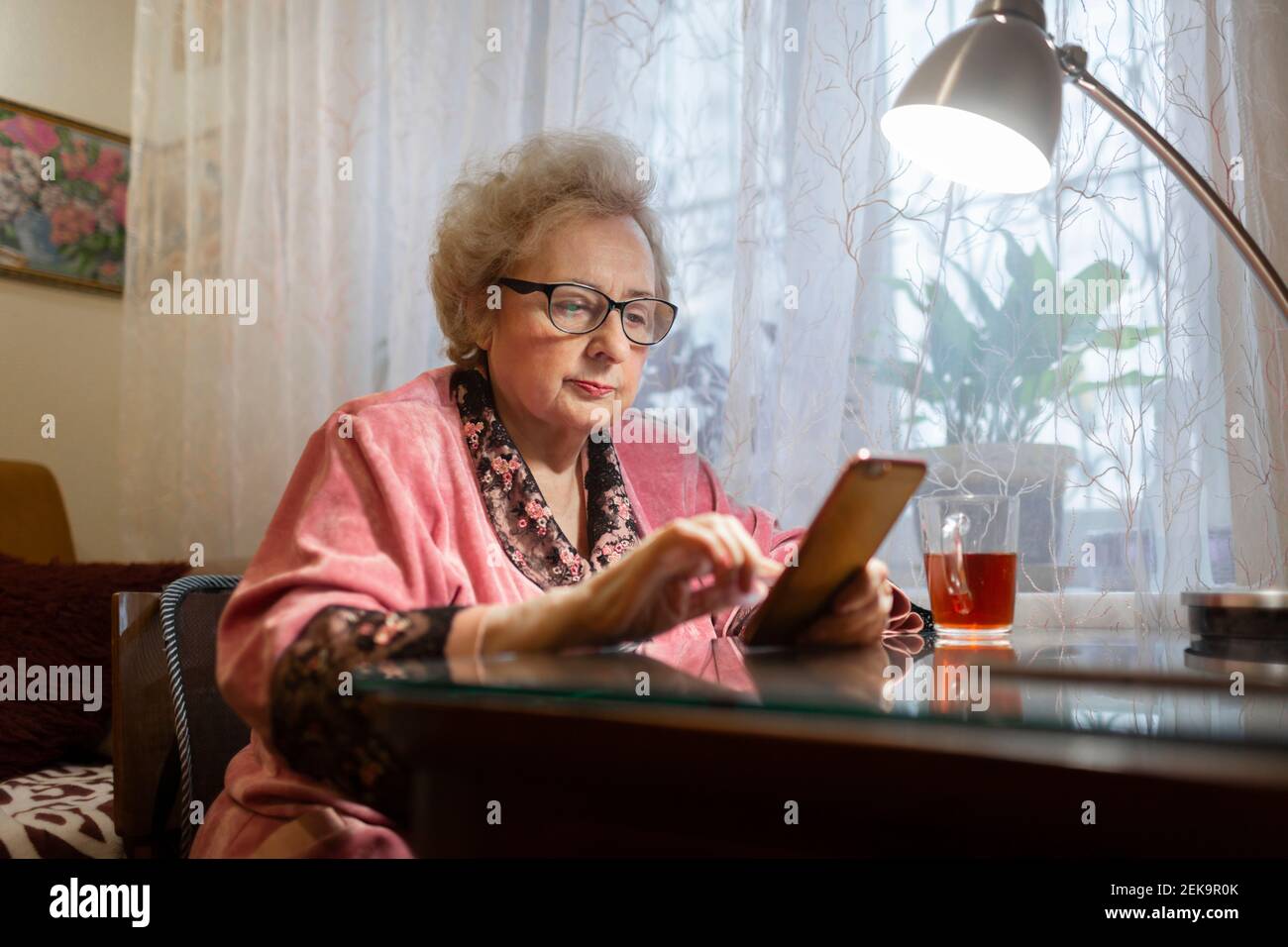 Femme âgée utilisant un smartphone sous une lampe électrique sur une table dans la salle de séjour Banque D'Images