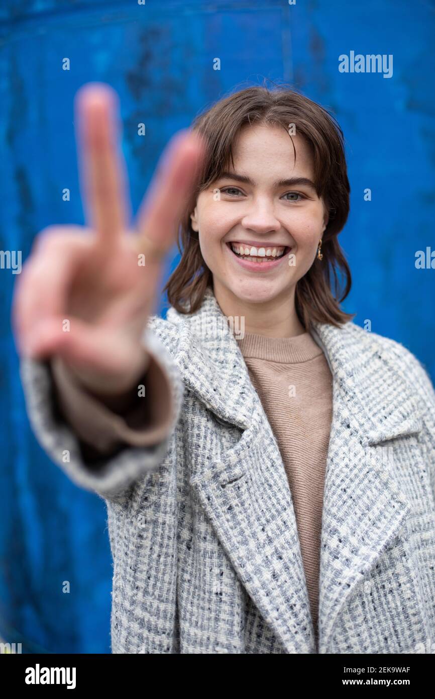 Femme souriante montrant un geste de paix contre un mur bleu Banque D'Images