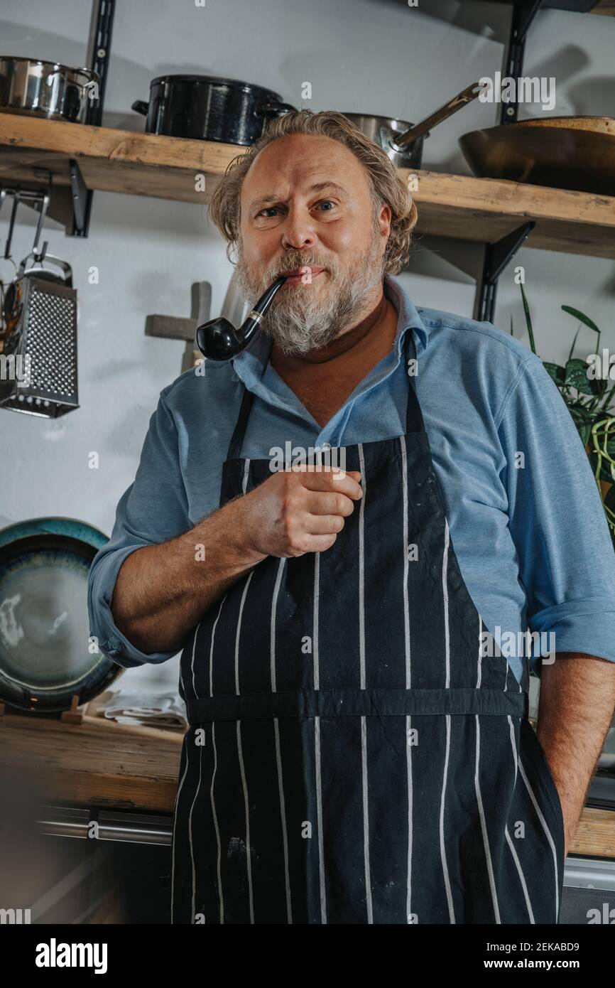 Chef mature portant un tablier debout avec le tuyau et les mains poches dans la cuisine Banque D'Images