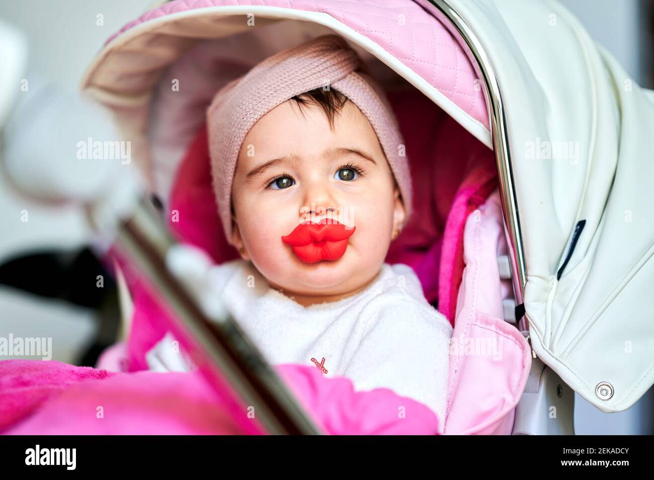 Bébé fille avec sucette en forme de lèvre regardant loin tout en étant assis poussette de bébé à la maison Banque D'Images