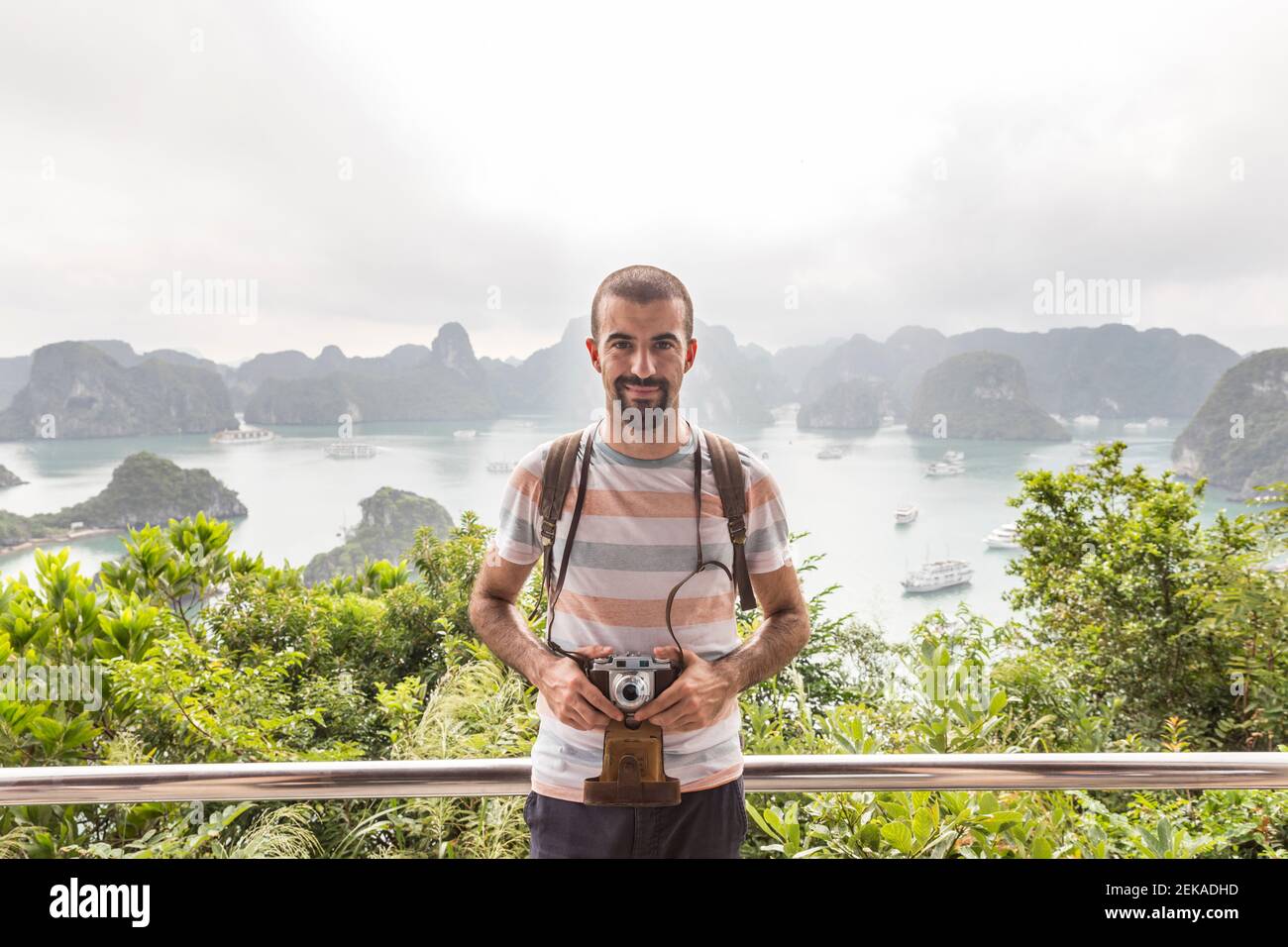 Homme souriant avec caméra d'époque debout sur le point d'observation contre la baie de Halong, Vietnam Banque D'Images
