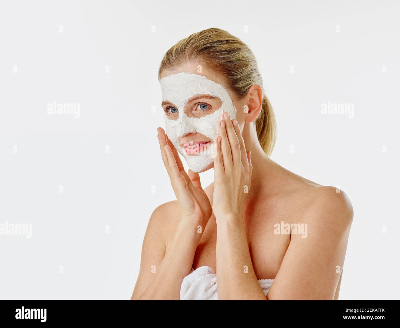 Femme souriante enveloppée d'une serviette appliquant un masque facial Banque D'Images