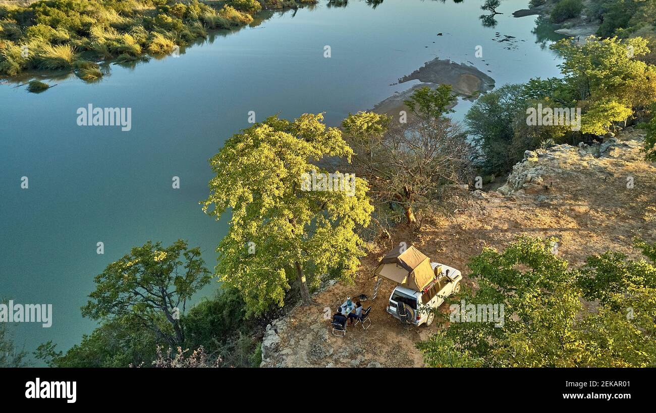Vue aérienne d'un couple campé avec leur jeep et tente sur le toit et prenant le petit déjeuner en face de la rivière, région de la rivière Cunene, Angola Banque D'Images