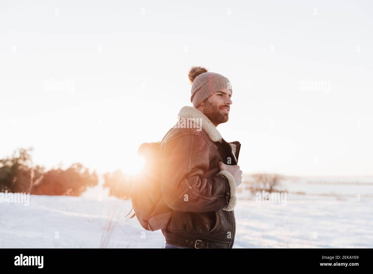 Jeune homme avec sac à dos qui regarde loin en se tenant dans la neige au coucher du soleil Banque D'Images