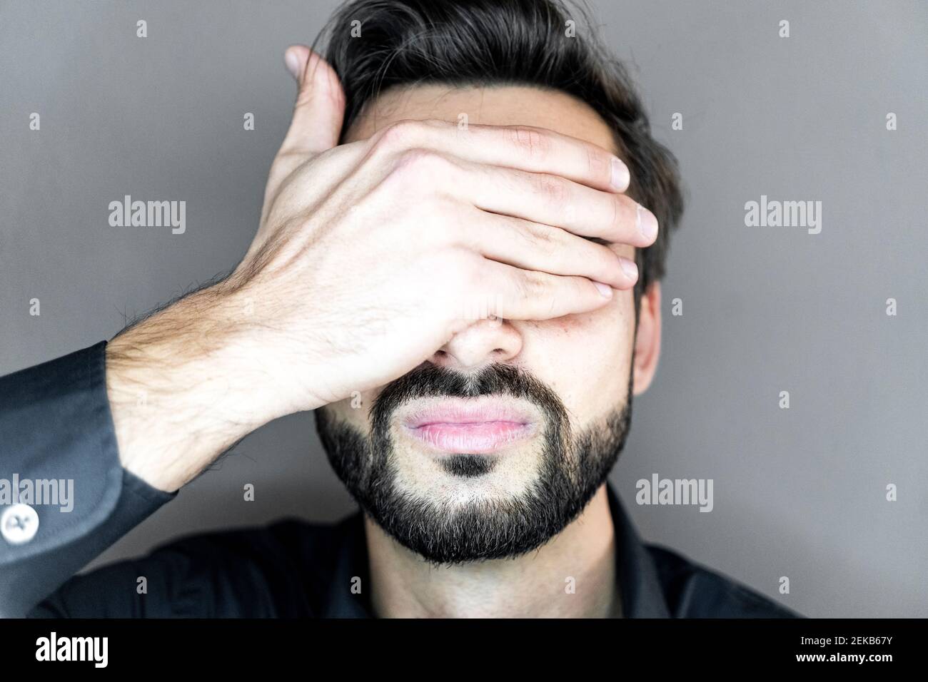 Jeune homme couvrant les yeux avec les mains contre le mur gris Banque D'Images