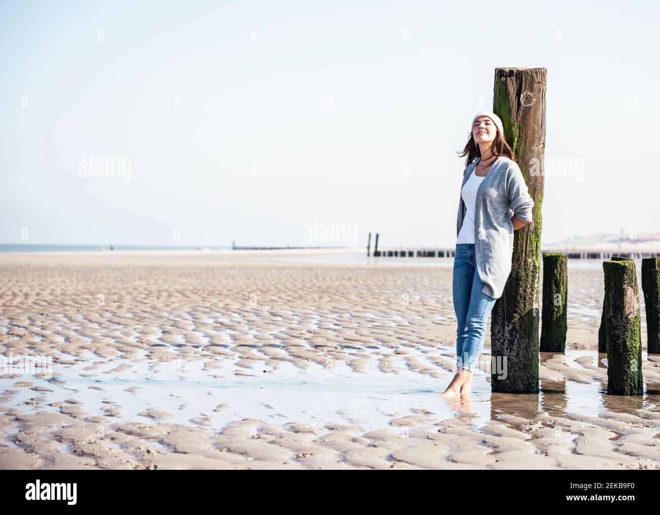 Jeune femme avec les yeux fermés, penchée sur un poteau en bois à plage Banque D'Images