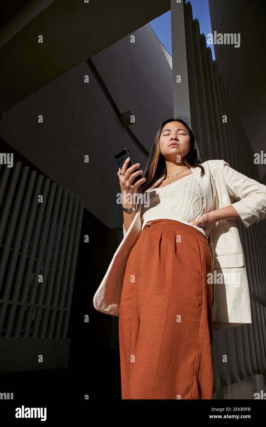 Jeune femme tenant un smartphone debout à la main hanche par jour ensoleillé Banque D'Images