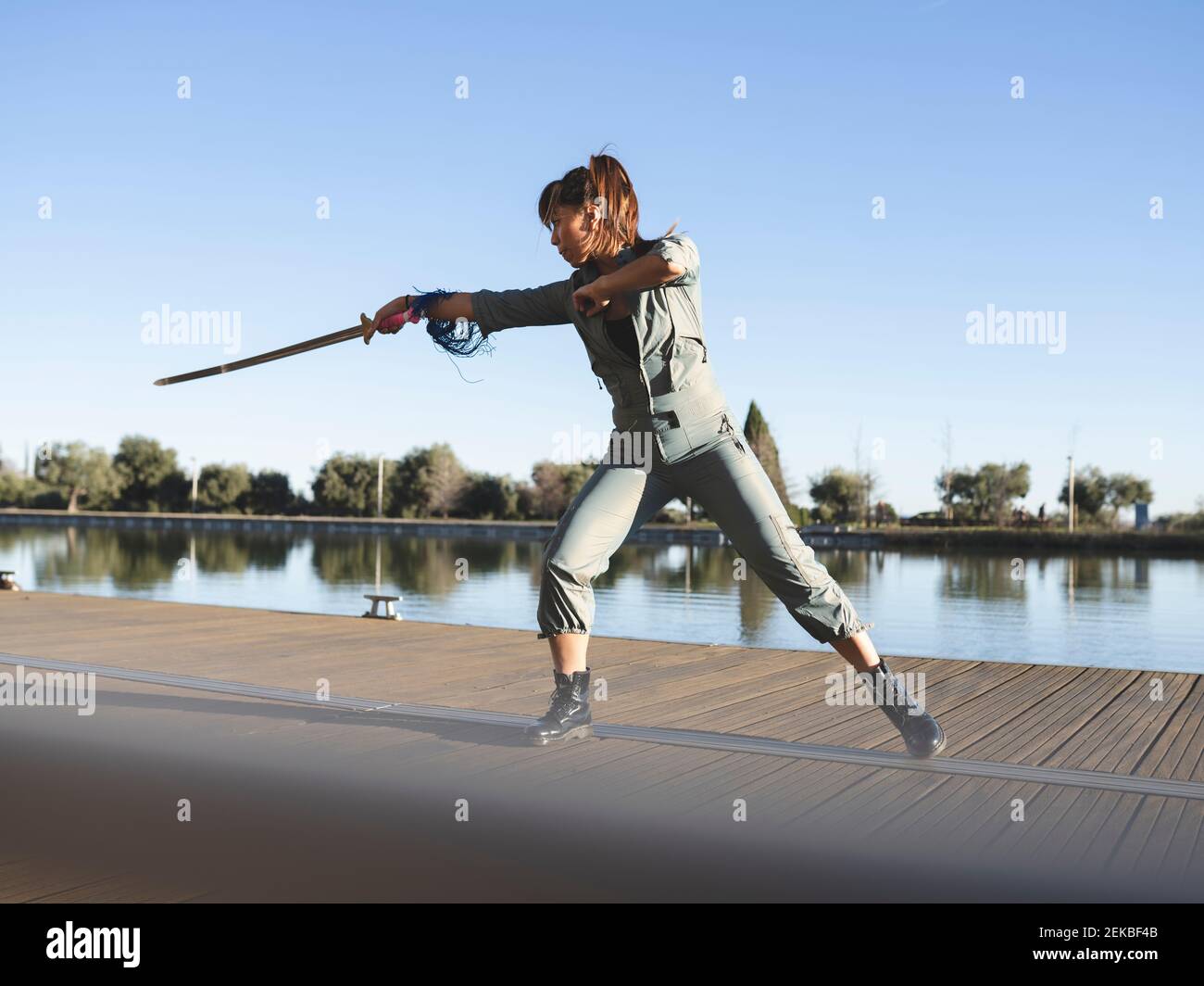 Jeune femme pratiquant les arts martiaux avec l'épée dans le parc contre lac par beau temps Banque D'Images Jeune femme pratiquant les arts martiaux avec l'épée dans le parc contre lac par beau temps Banque D'Images