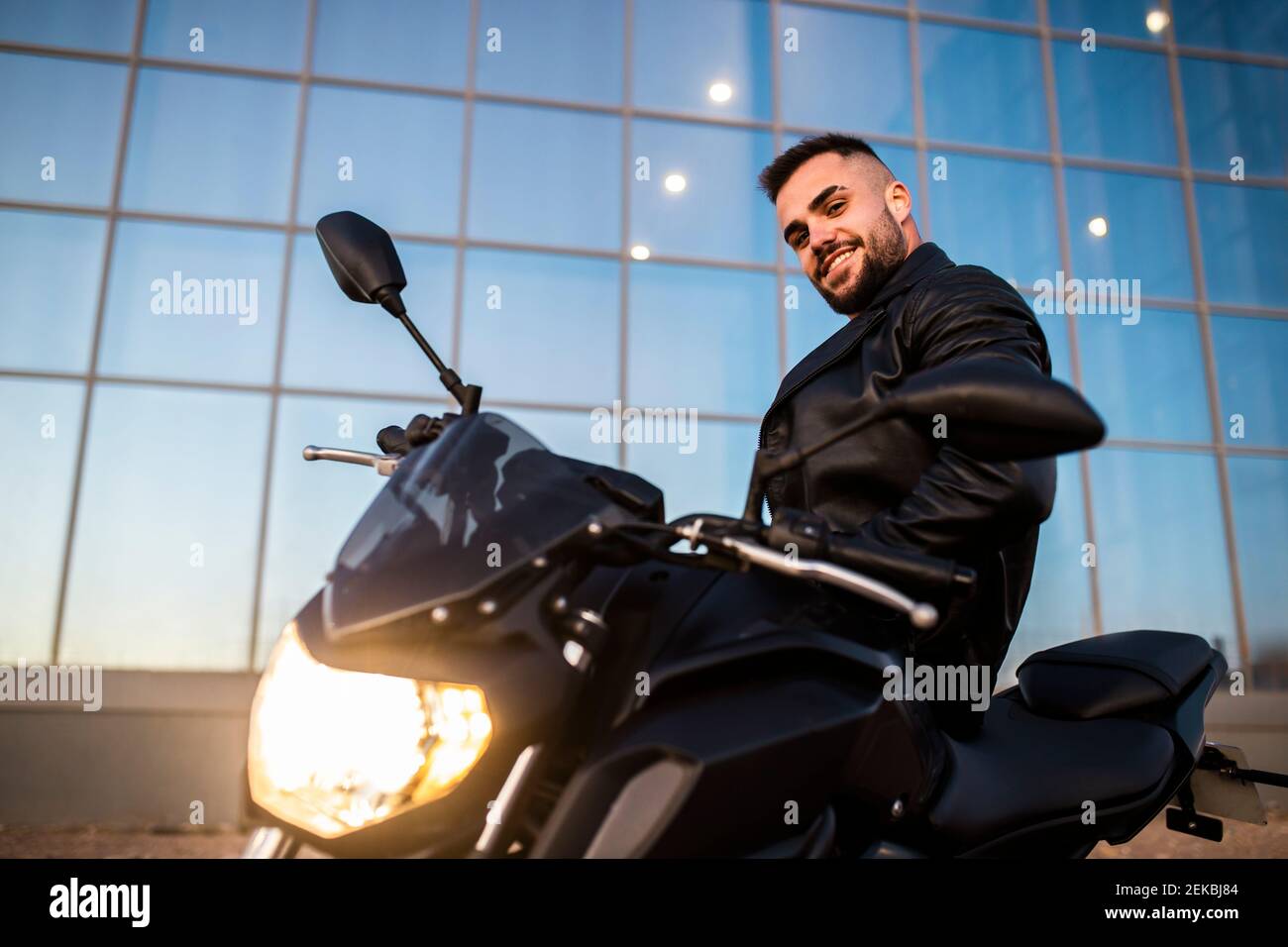 Homme souriant en cuir veste penchée sur la moto par moderne bâtiment Banque D'Images