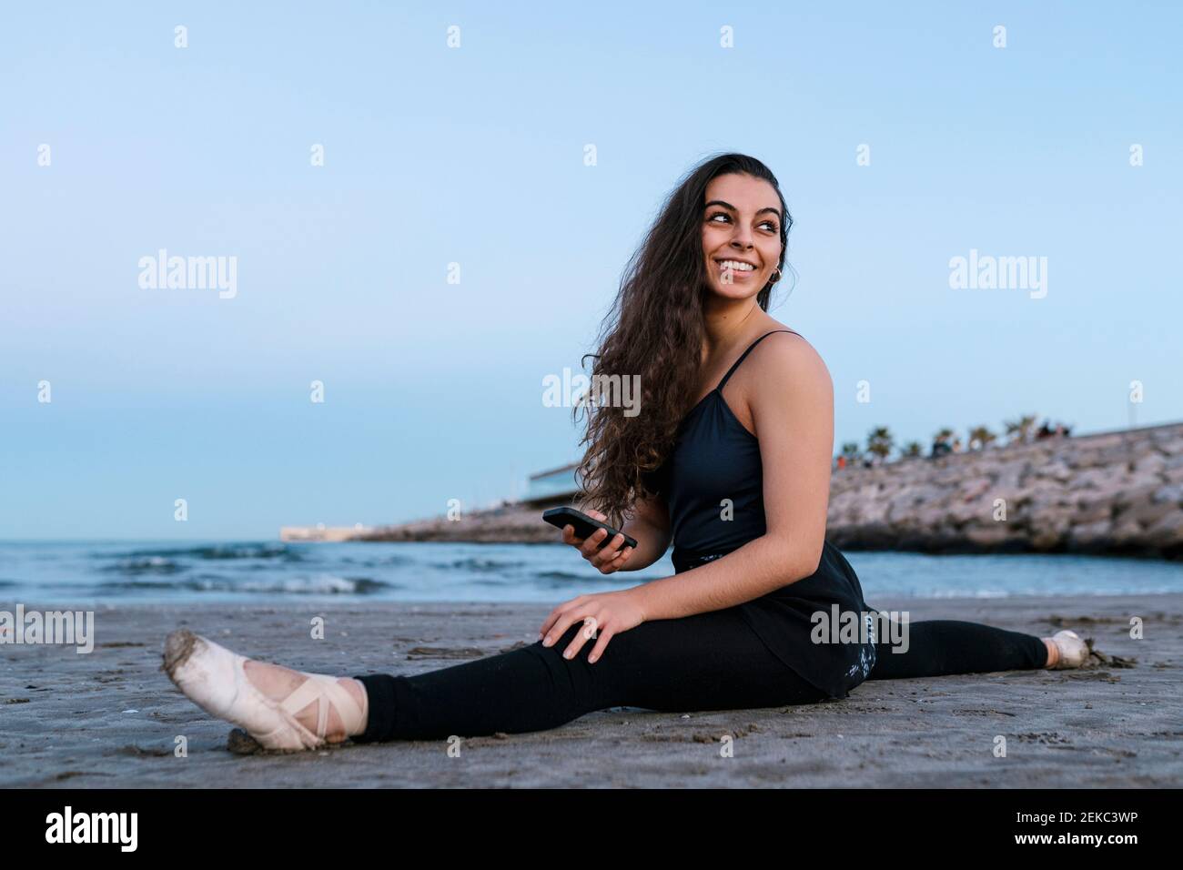 Danseuse de ballet souriante regardant l'épaule tout en faisant la fente de jambe à la plage contre un ciel dégagé Banque D'Images