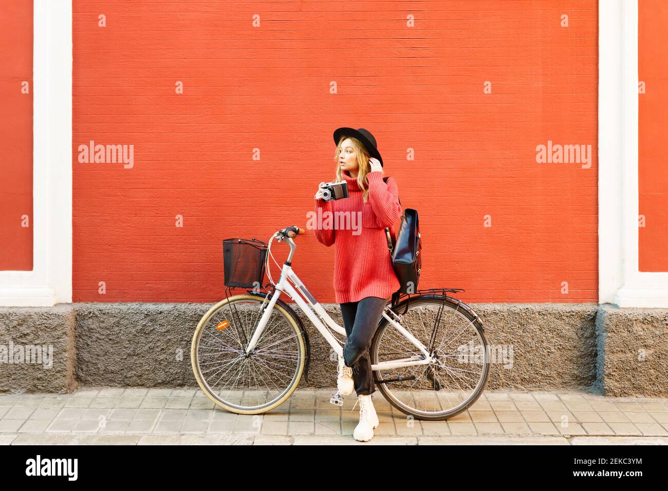 Jeune femme portant un chapeau tenant un appareil photo vintage tout en se tenant debout avec un vélo contre le mur rouge Banque D'Images