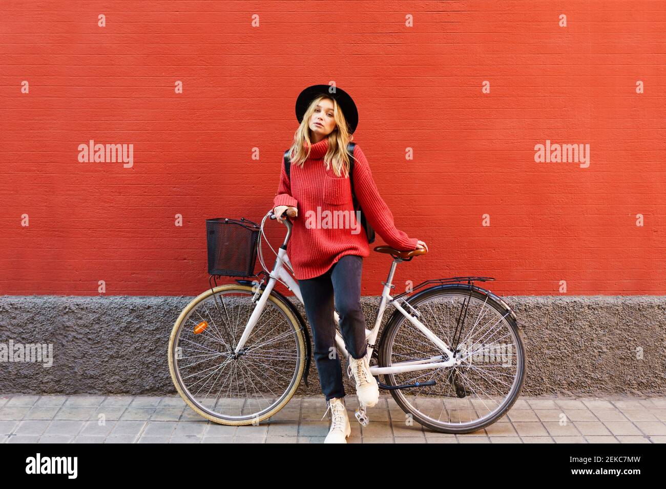 Jeune femme blonde portant un chapeau debout avec un vélo contre un mur rouge Banque D'Images