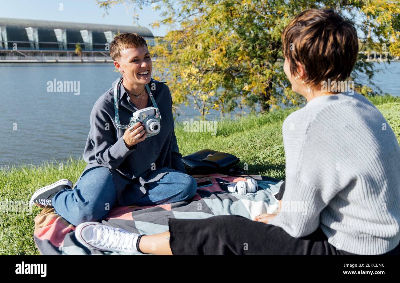 Femme souriante avec un appareil photo regardant un ami assis contre rivière au parc Banque D'Images