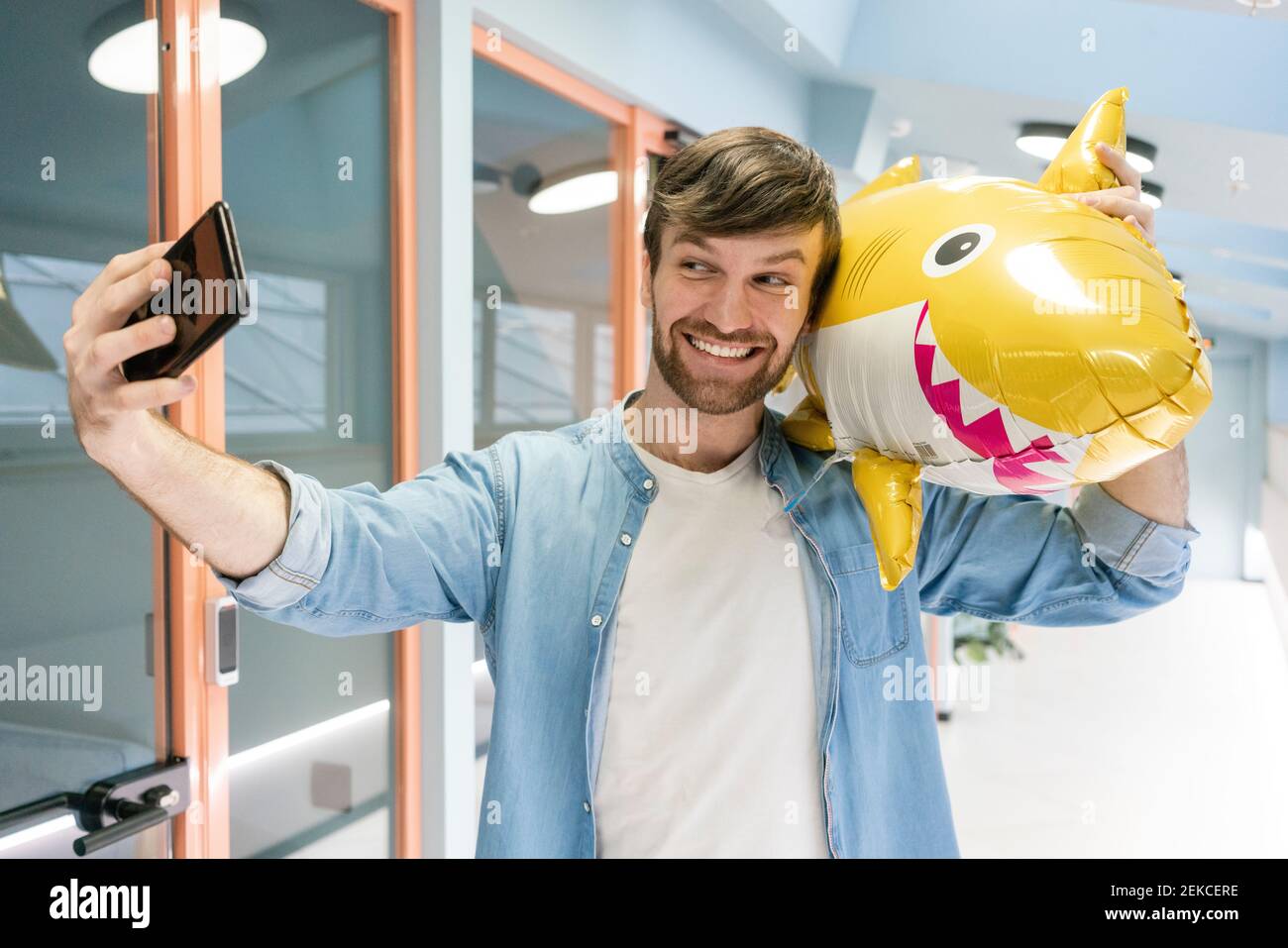 Homme d'affaires souriant et créatif prenant un selfie avec un requin-montgolfière dans le couloir sur le lieu de travail Banque D'Images