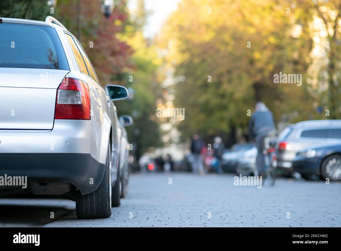 Voitures garées dans une rangée sur le côté de la rue de la ville le jour d'automne lumineux avec des personnes floues marchant sur la zone piétonne. Banque D'Images