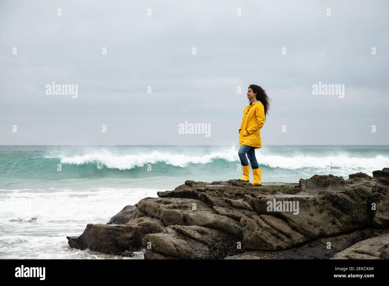 Jeune femme en imperméable jaune regardant la vue sur la mer pendant debout sur le rocher Banque D'Images