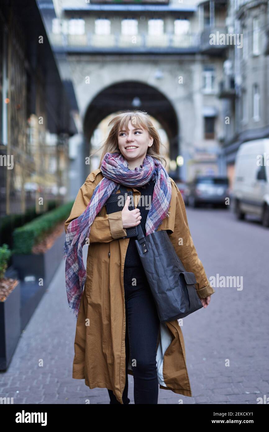 Photo de style urbain d'une femme blonde élégante portant un long manteau  de tranchée orange, un Jean noir et une écharpe avec sac noir zéro déchet,  marchant dans la rue de l'Euro