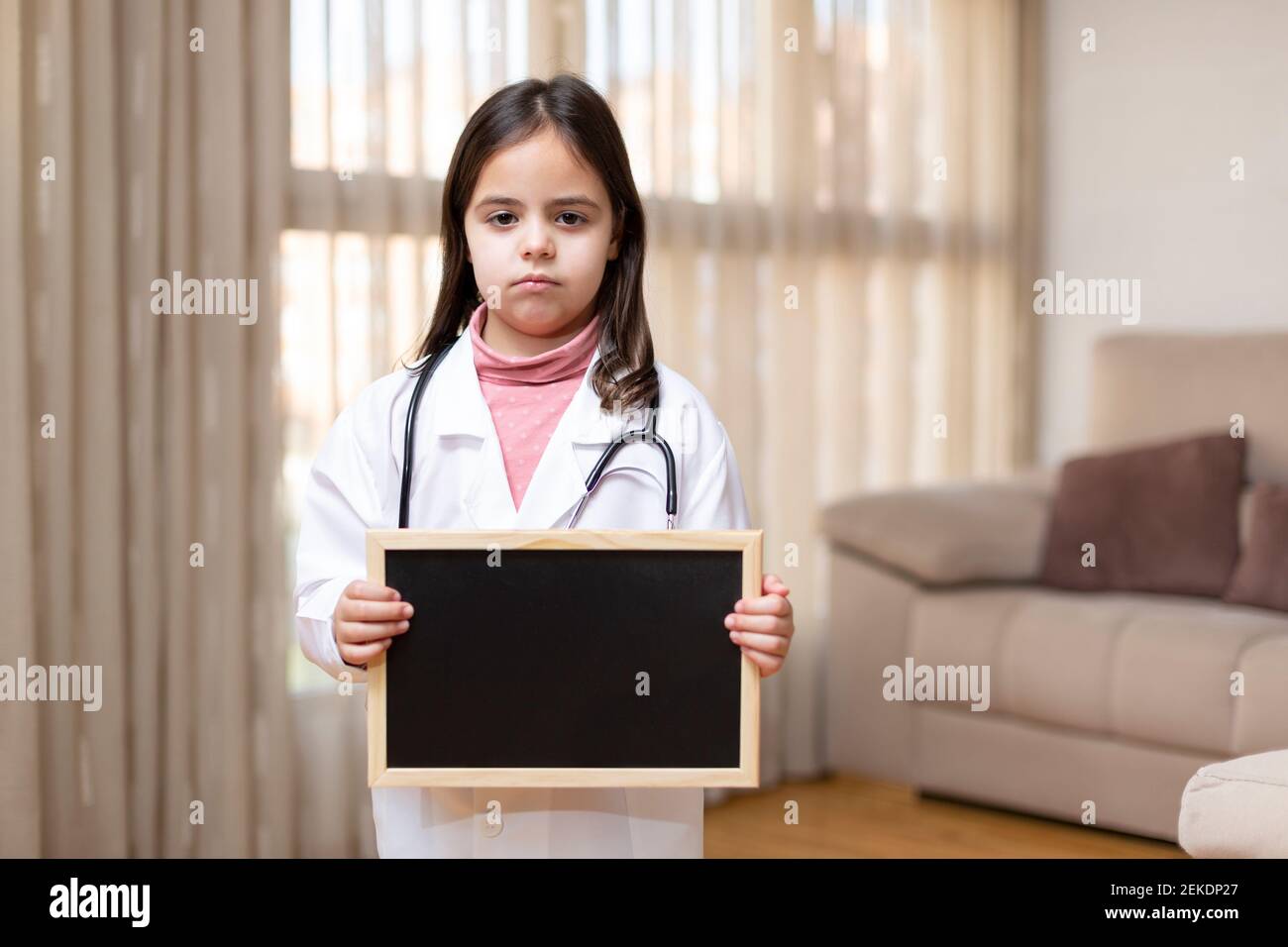 Petit enfant habillé comme un médecin et avec un geste sérieux tenant un tableau noir à la maison. Espace pour le texte. Banque D'Images