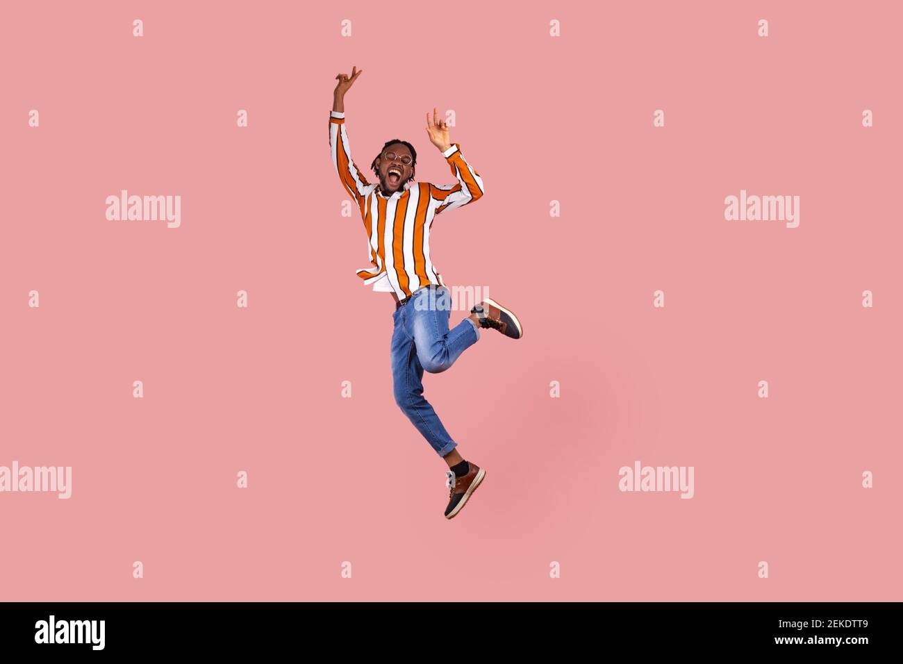Homme afro-américain écostatique plein d'enthousiasme avec des dreadlocks dans une chemise rayée, qui sautant fortement, montrant le geste de la victoire, célébrant son tripump Banque D'Images