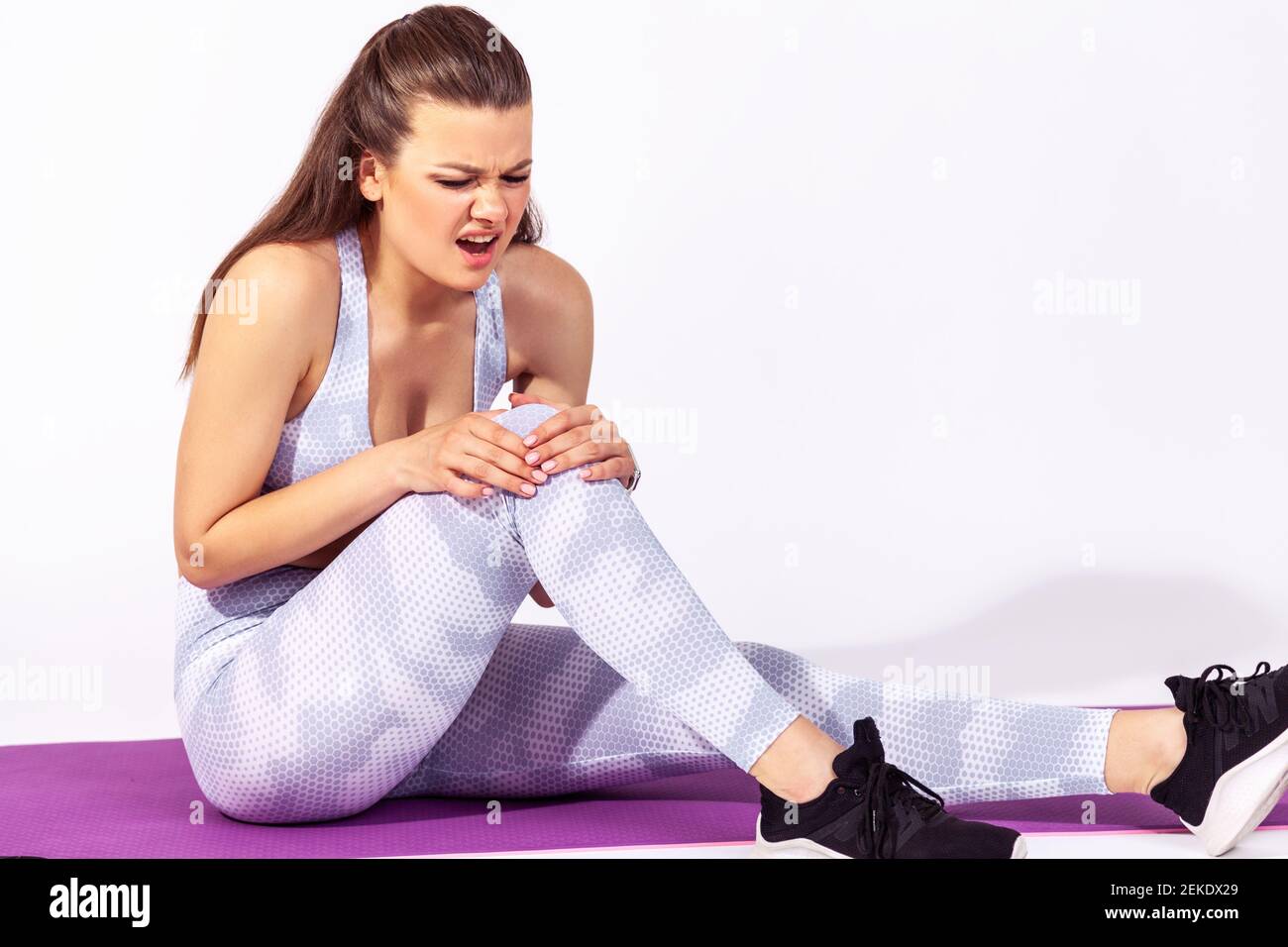 Femme d'athlétisme en vêtements de sport blancs hurlant au genou, assis sur un tapis de gym, souffrant de tension musculaire après l'entraînement de yoga. Studio intérieur Banque D'Images