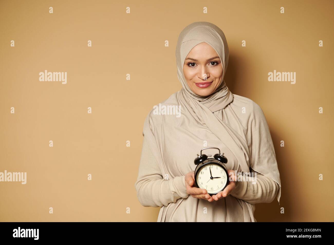 Jeune femme musulmane gaie dans le hijab tenant le réveil dans les mains et souriant mignon à la caméra debout sur un fond beige avec un espace de copie. Banque D'Images