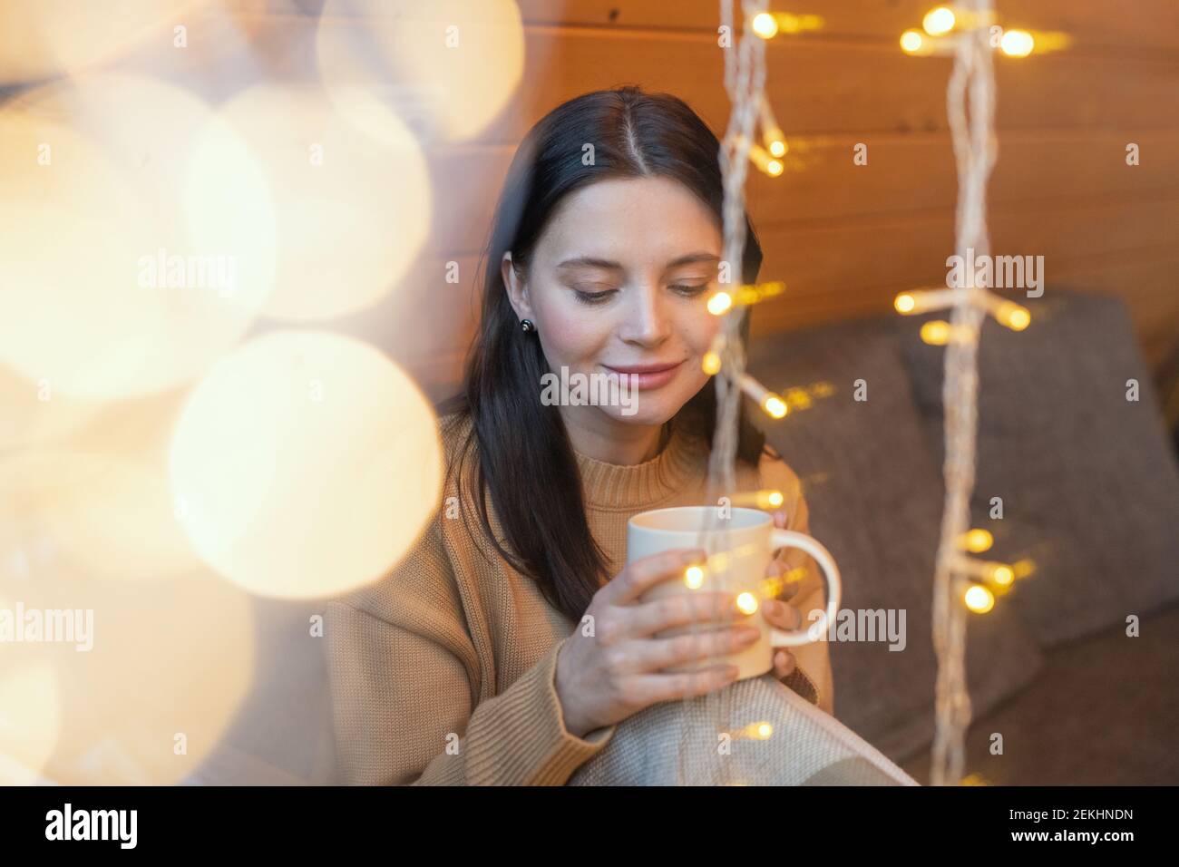 Jeune femme sereine brune avec une tasse de thé chaud assis sur le canapé par une grande fenêtre de la maison de campagne en hiver week-end et repos Banque D'Images