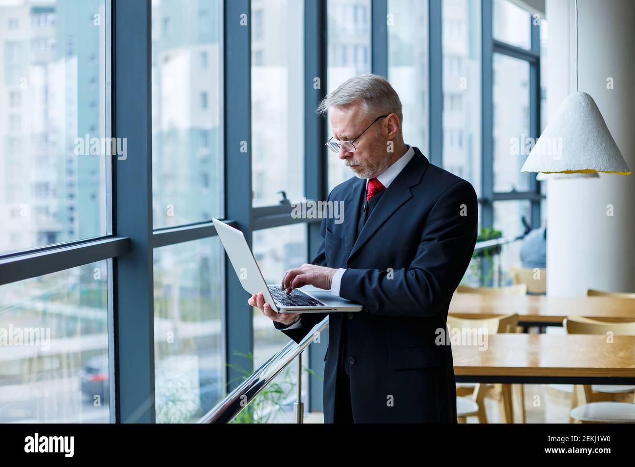 Sourire heureux directeur général pense à son développement de carrière réussi tout en se tenant debout avec un ordinateur portable dans son bureau près de l'arrière-plan d'une victoire Banque D'Images