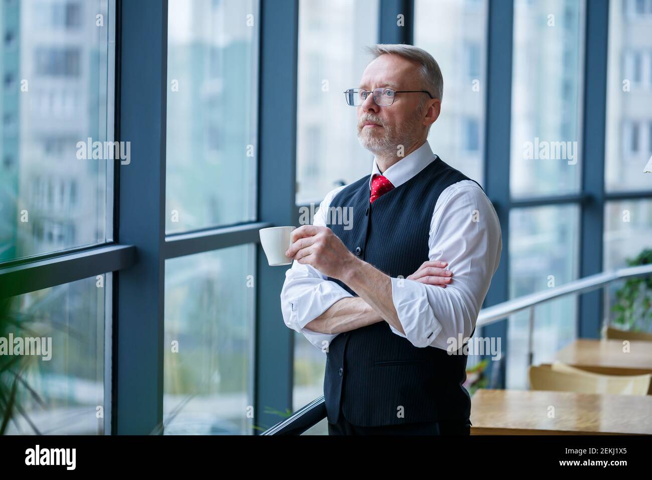 Sourire heureux directeur général pense à son développement de carrière réussi tout en se tenant avec une tasse de café dans sa main dans son bureau près de la Banque D'Images