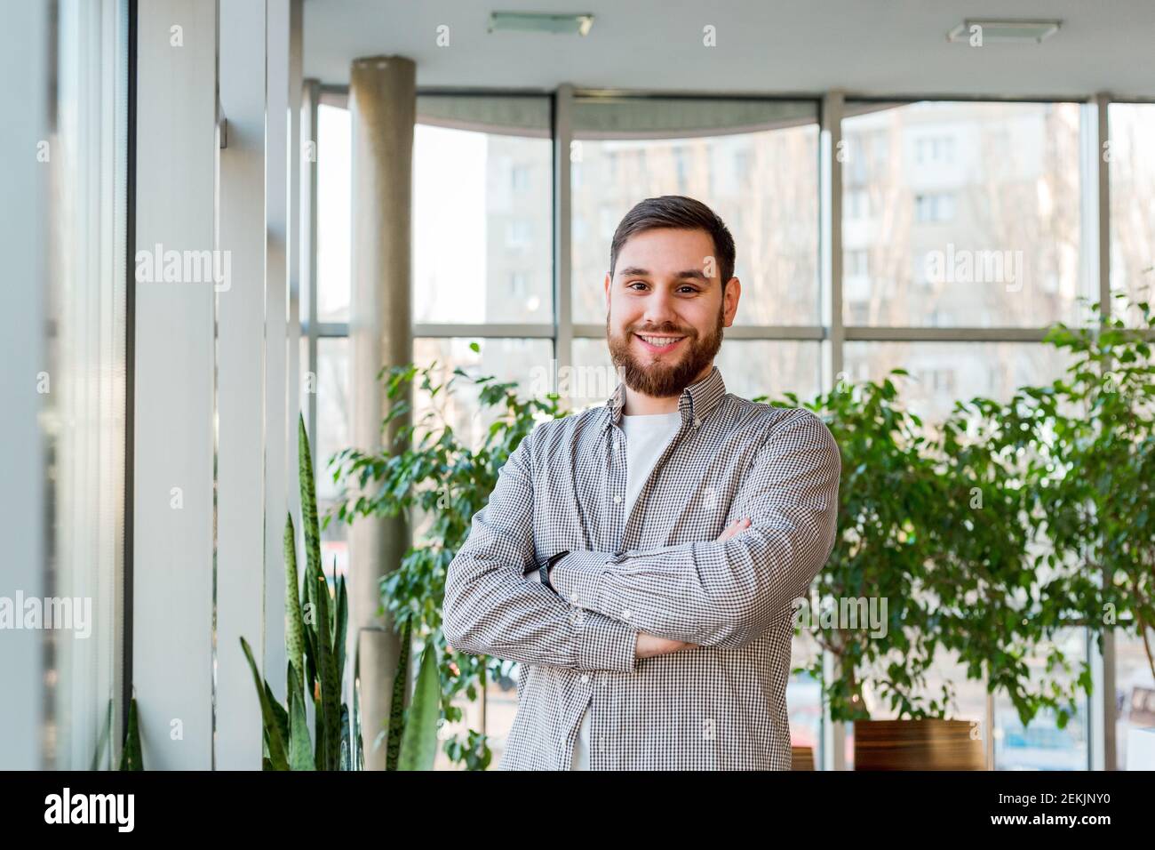 Homme caucasien souriant debout près de la fenêtre dans le bureau. Jeune homme intelligent barbu avec succès et bras croisés. Beau homme d'affaires dans une chemise décontractée. Gratuit Banque D'Images
