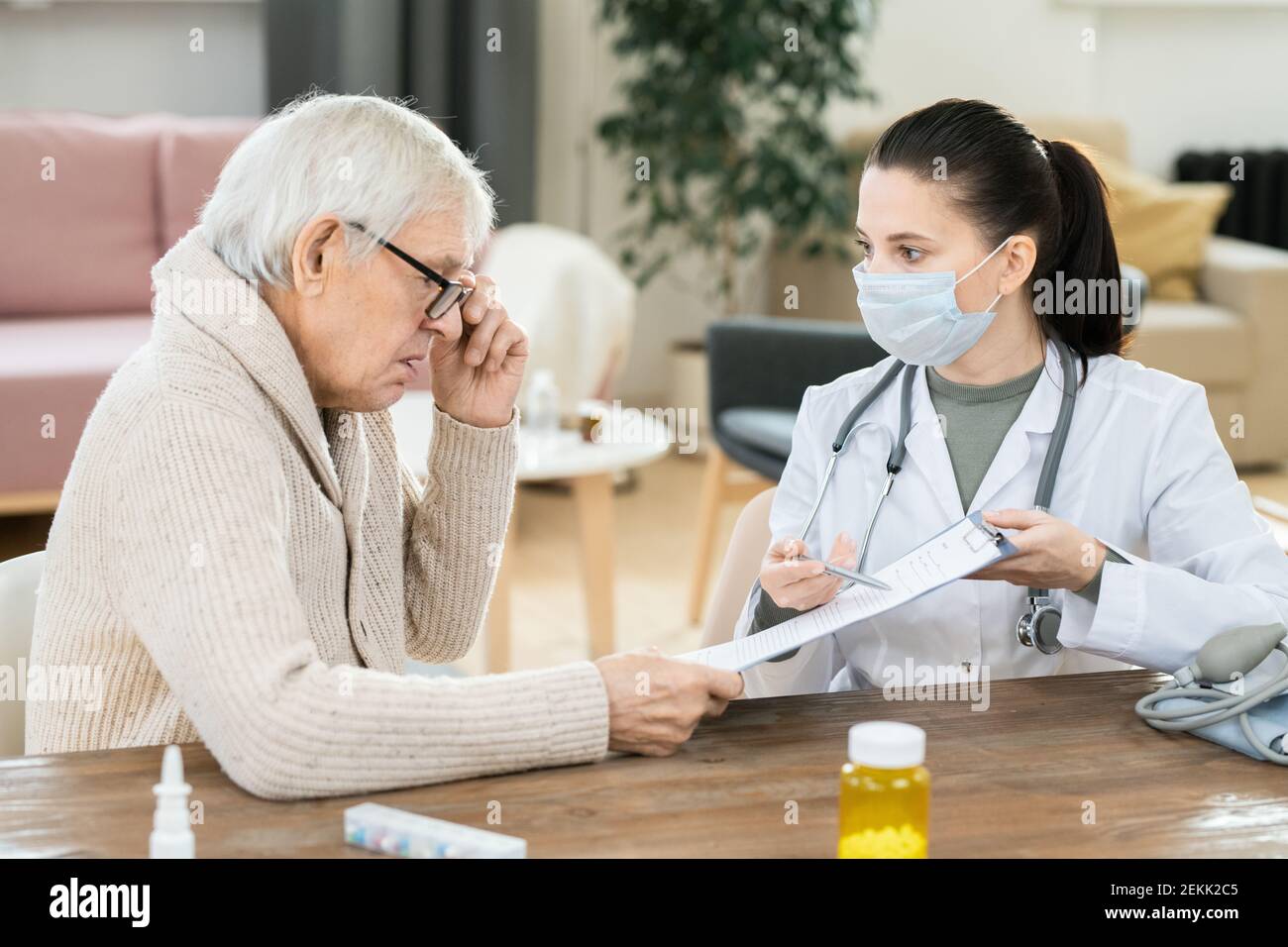 Homme âgé malade regardant par un document médical détenu par un jeune femme médecin assise à table devant lui pendant consultation à domicile Banque D'Images