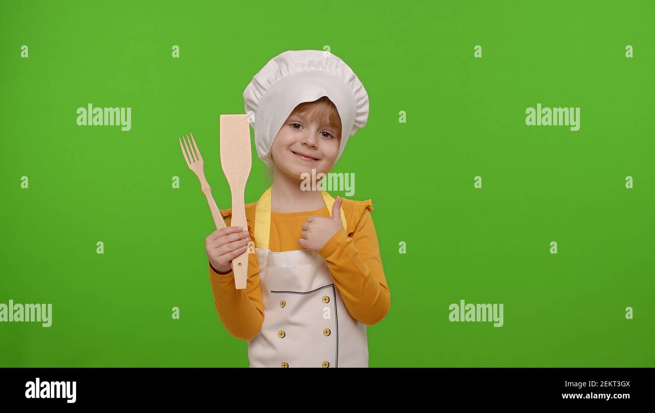 Drôle mignon caucasien jeune fille habillé comme cuisinier boulanger de chef dans un tablier et chapeau avec fourchette en bois et la spatule, sourire, les nodes tête en accord sur fond de clé de chroma. Pouce vers le haut Banque D'Images