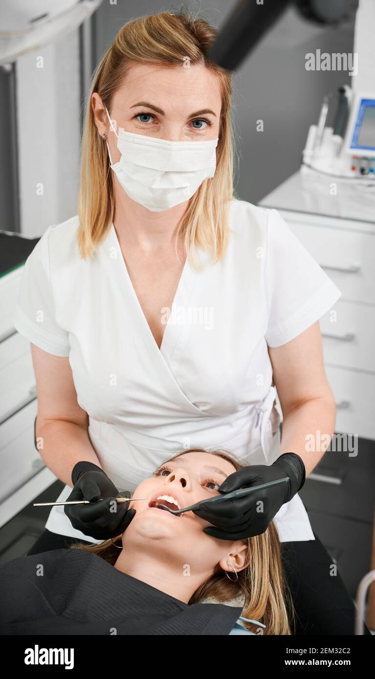 Docteur En Gants Médicaux Noirs Examine Les Dents De La Jeune Fille En  Utilisant Un Microscope Dentaire Et Un Miroir Dans Le Bureau Du Dentiste.  La Femme A Un Bavoir Du Patient